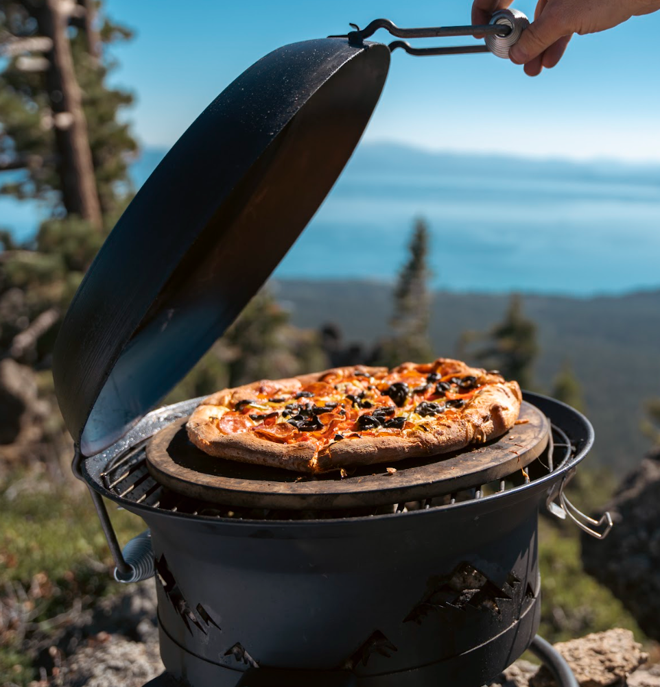 4-in-1 portable propane camping grill shown in pizza oven mode with pizza stone heating up pizza in the backcountry